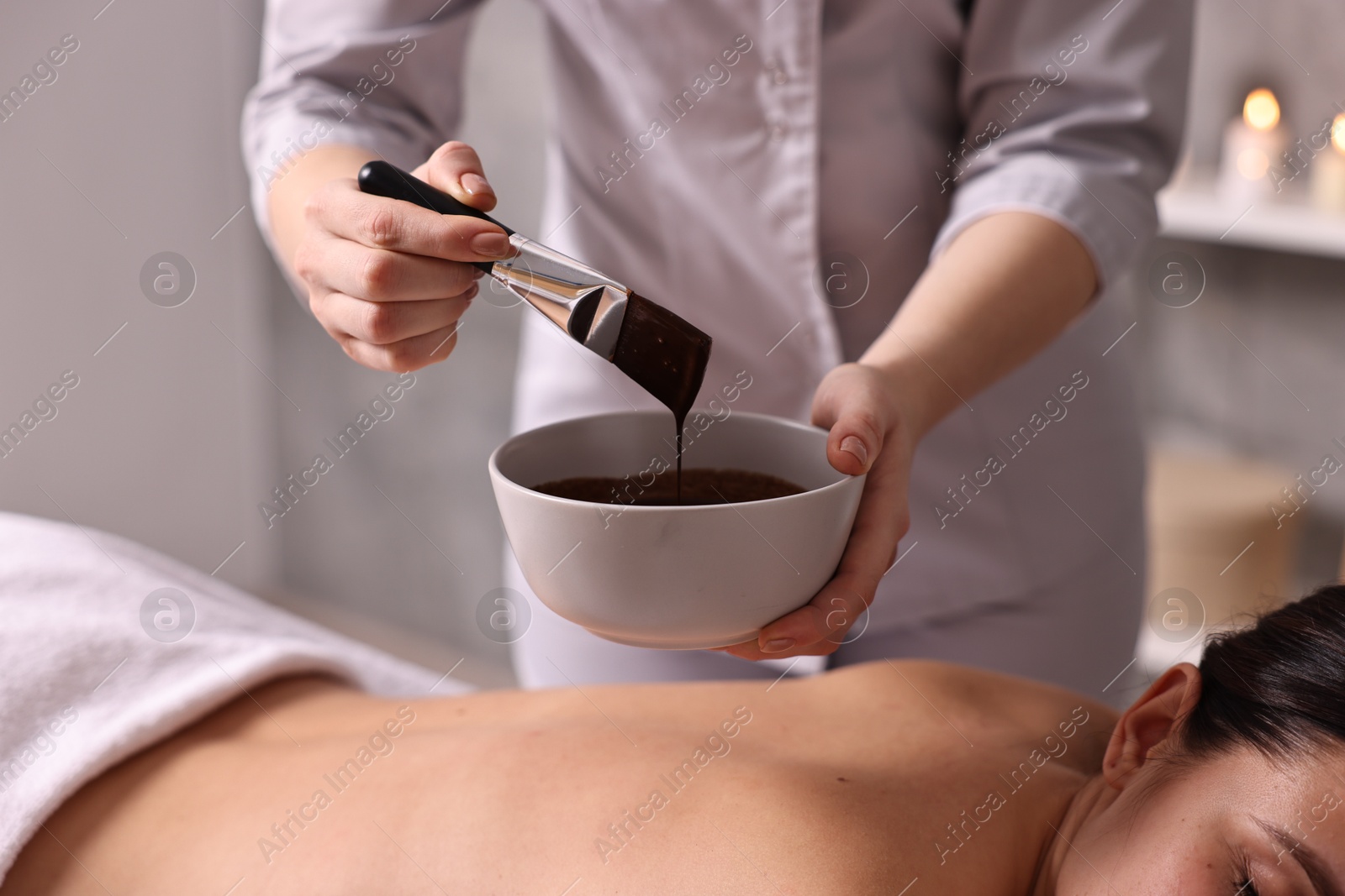 Photo of Spa worker with cosmetic product for chocolate body wrap near woman in salon, closeup