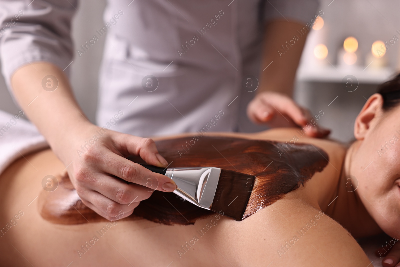 Photo of Chocolate body wrap. Spa worker applying mask onto woman's back in salon, closeup