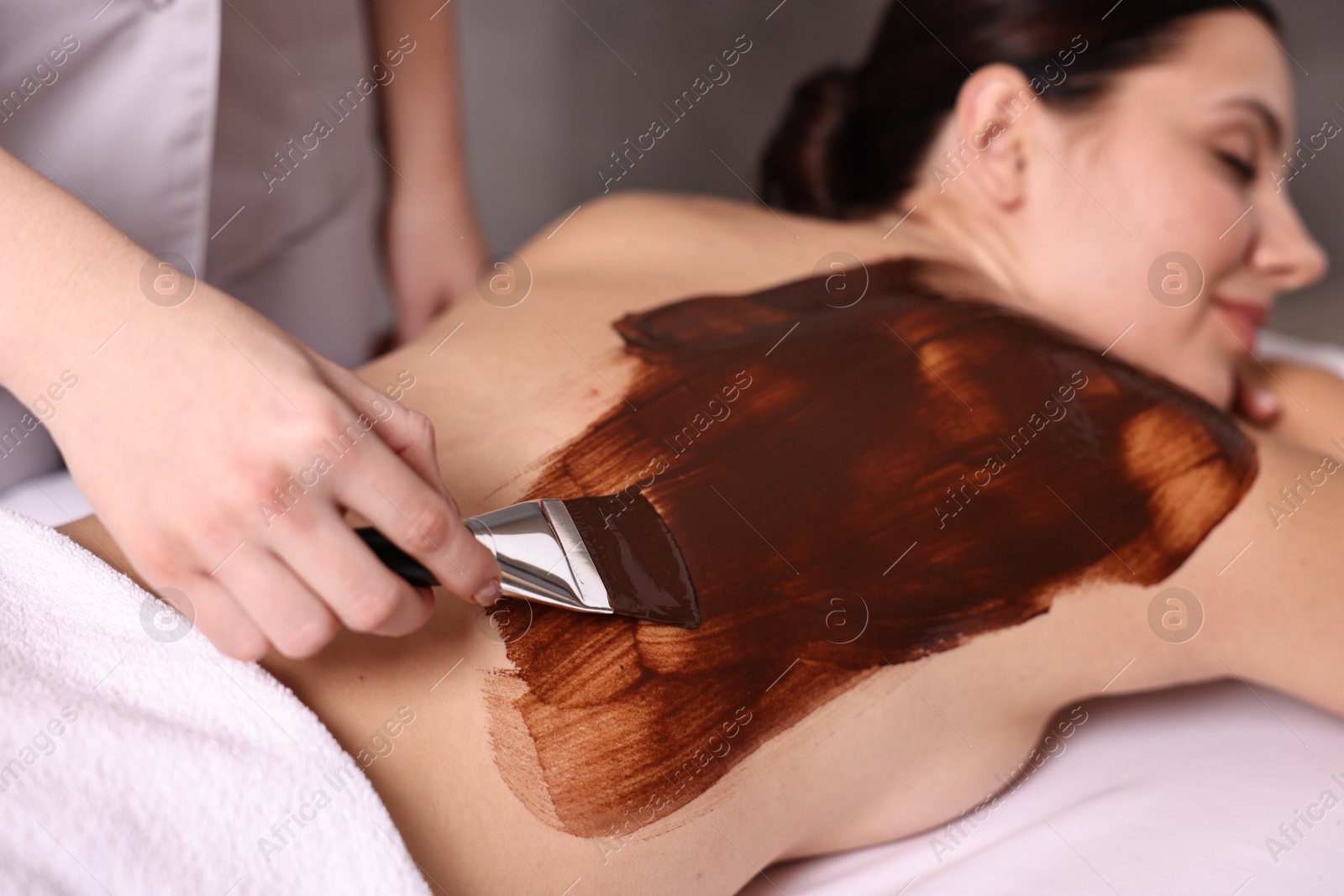 Photo of Chocolate body wrap. Spa worker applying mask onto woman's back in salon, closeup