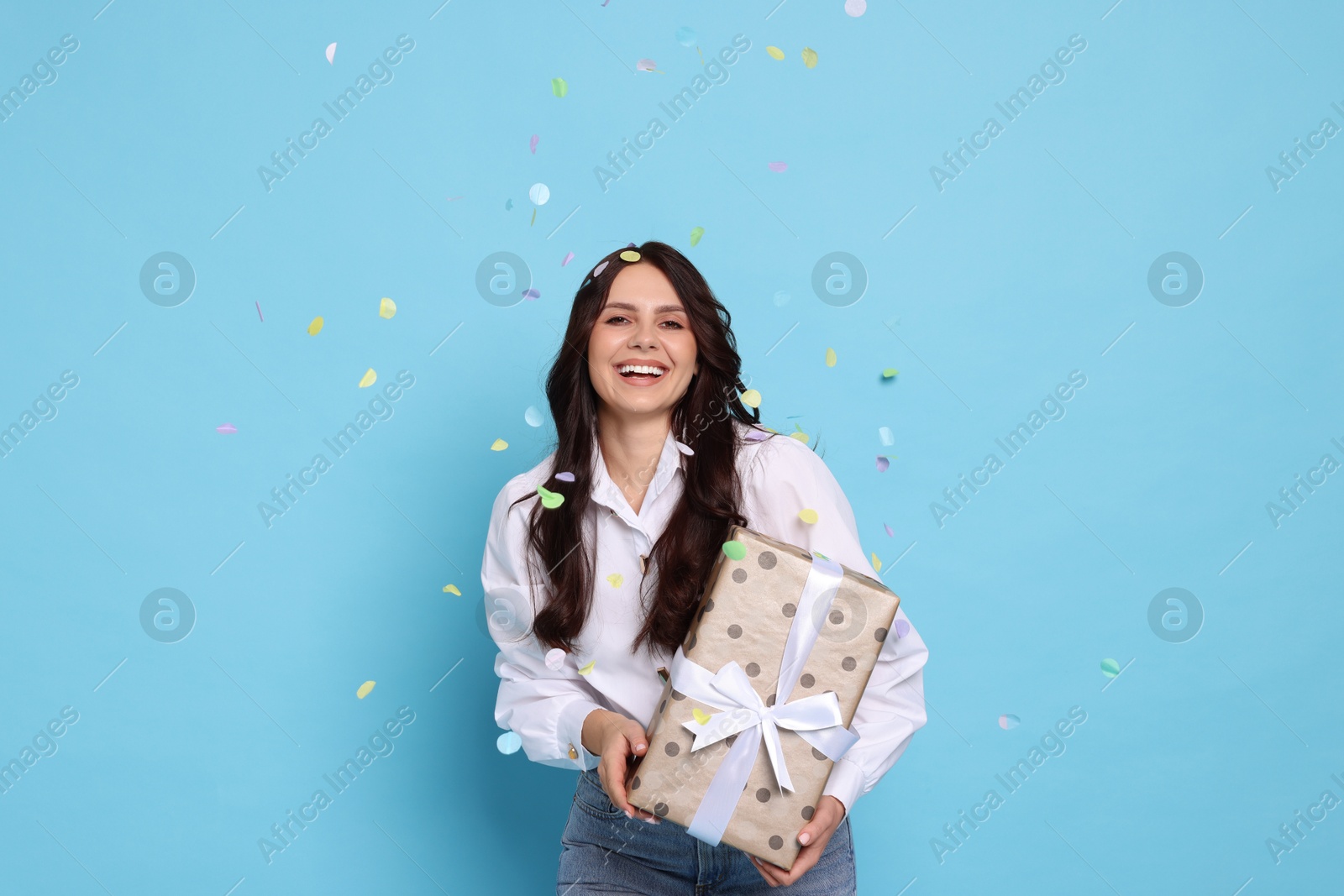 Photo of Happy woman with gift box under falling confetti on light blue background. Surprise party