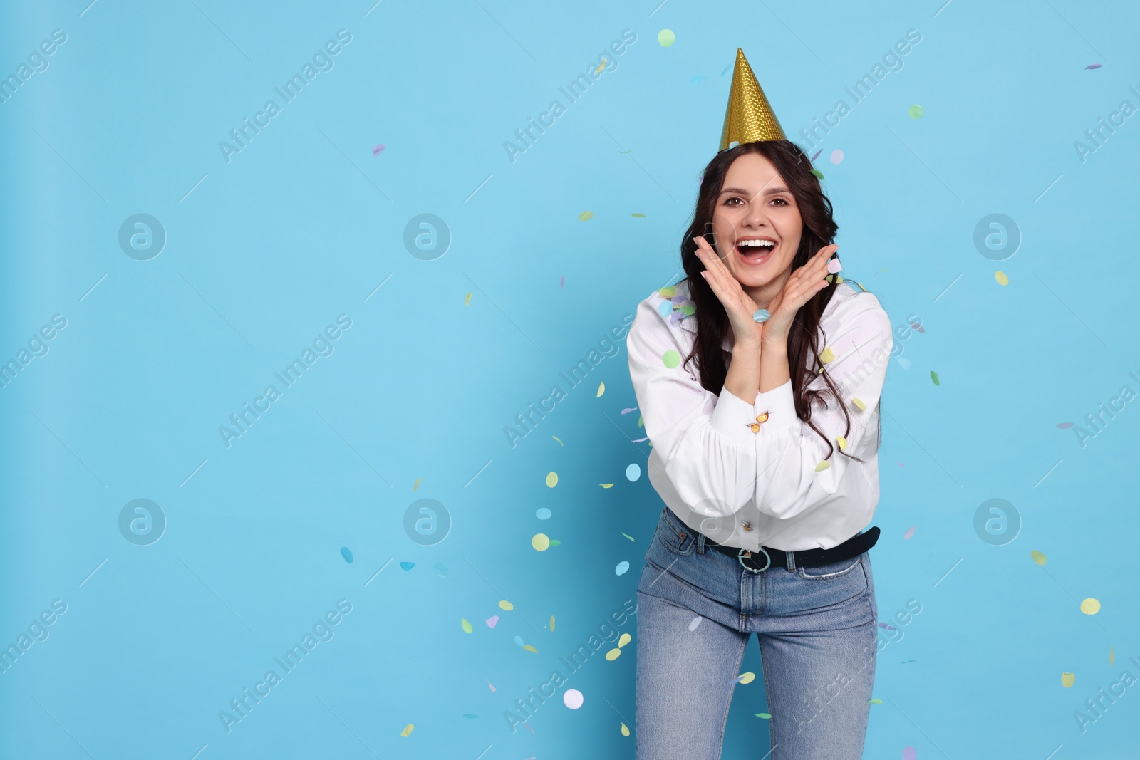 Photo of Happy woman in conical paper hat under falling confetti on light blue background, space for text. Surprise party