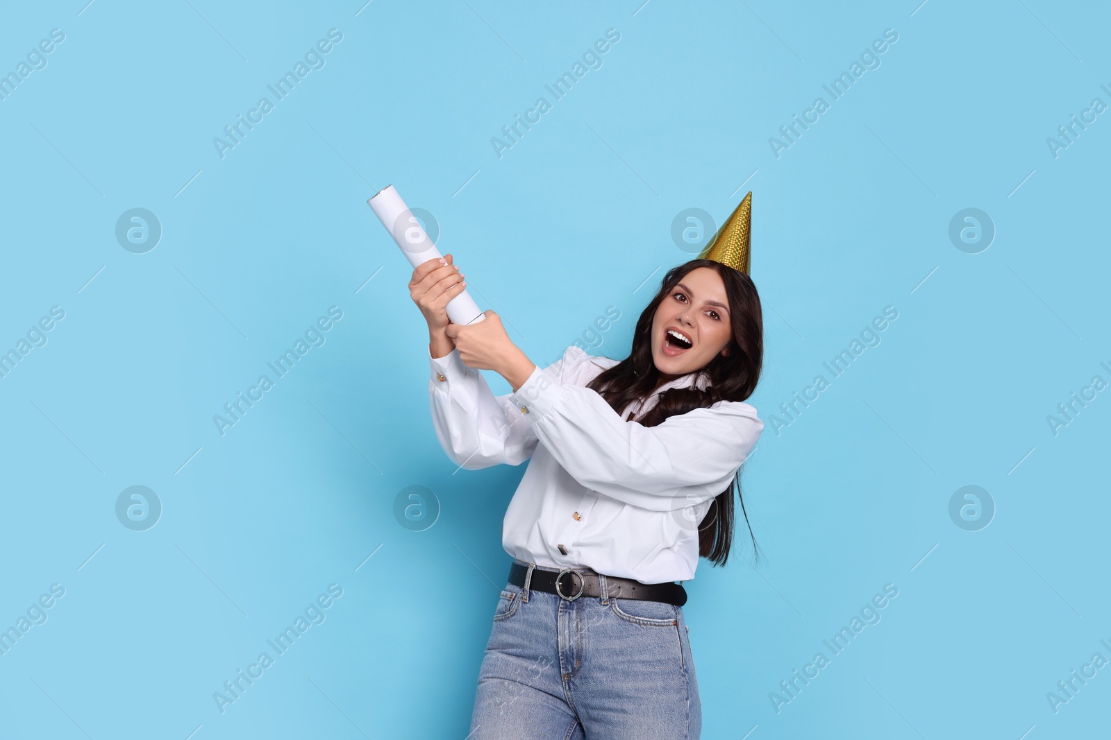 Photo of Happy woman in conical paper hat blowing up confetti popper on light blue background. Surprise party