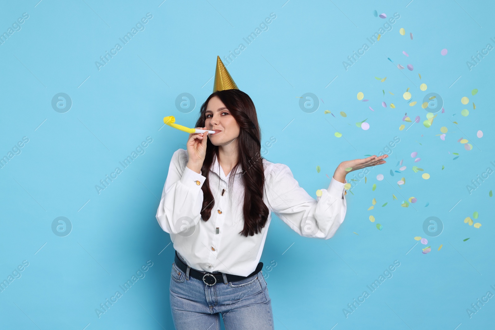 Photo of Woman in conical paper hat with blower and flying confetti on light blue background. Surprise party