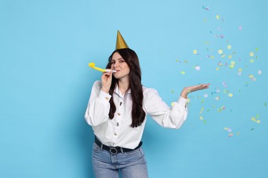 Woman in conical paper hat with blower and flying confetti on light blue background. Surprise party