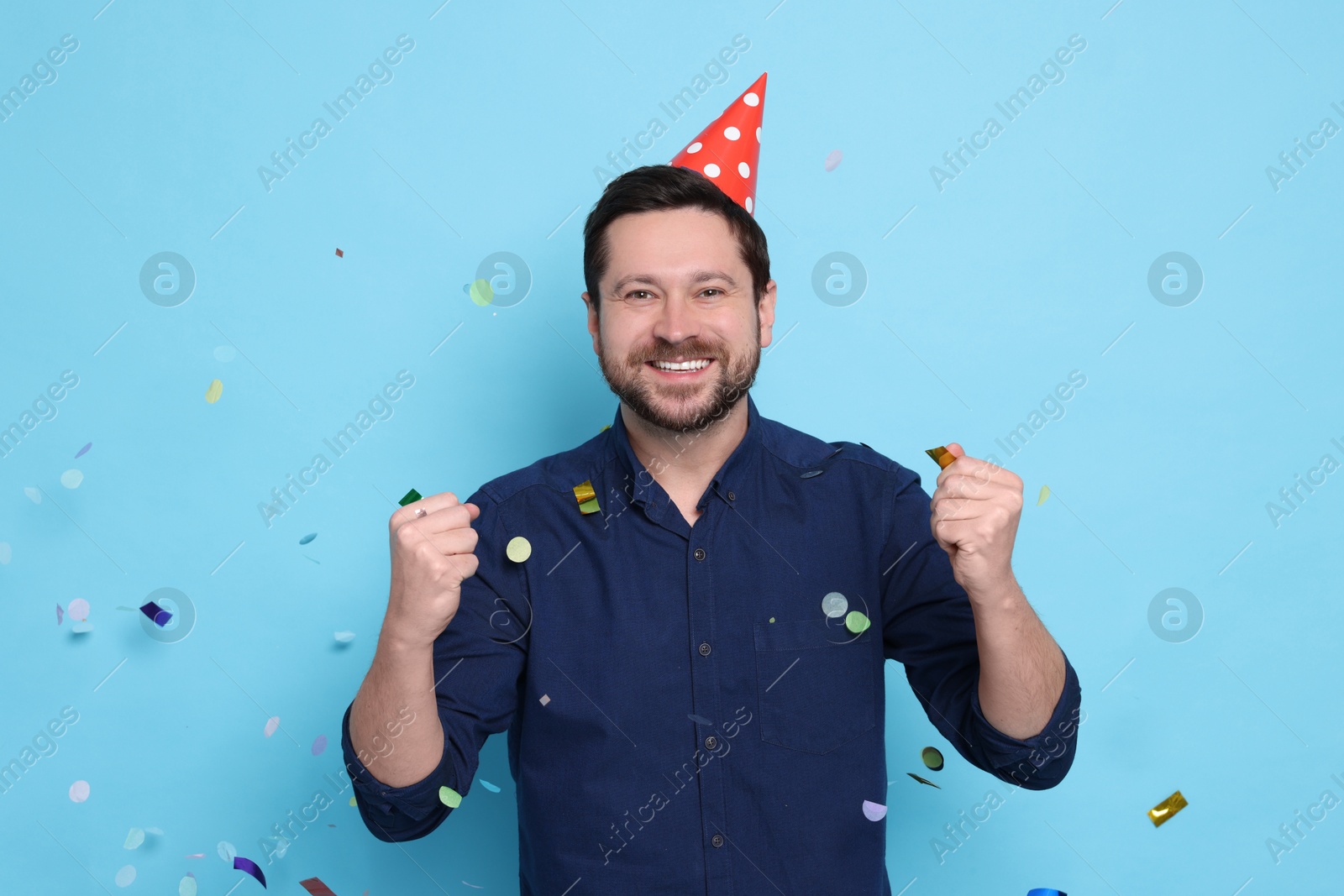 Photo of Happy man in conical paper hat and flying confetti on light blue background. Surprise party