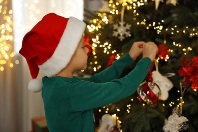 Photo of Cute little boy in Santa hat decorating Christmas tree at home