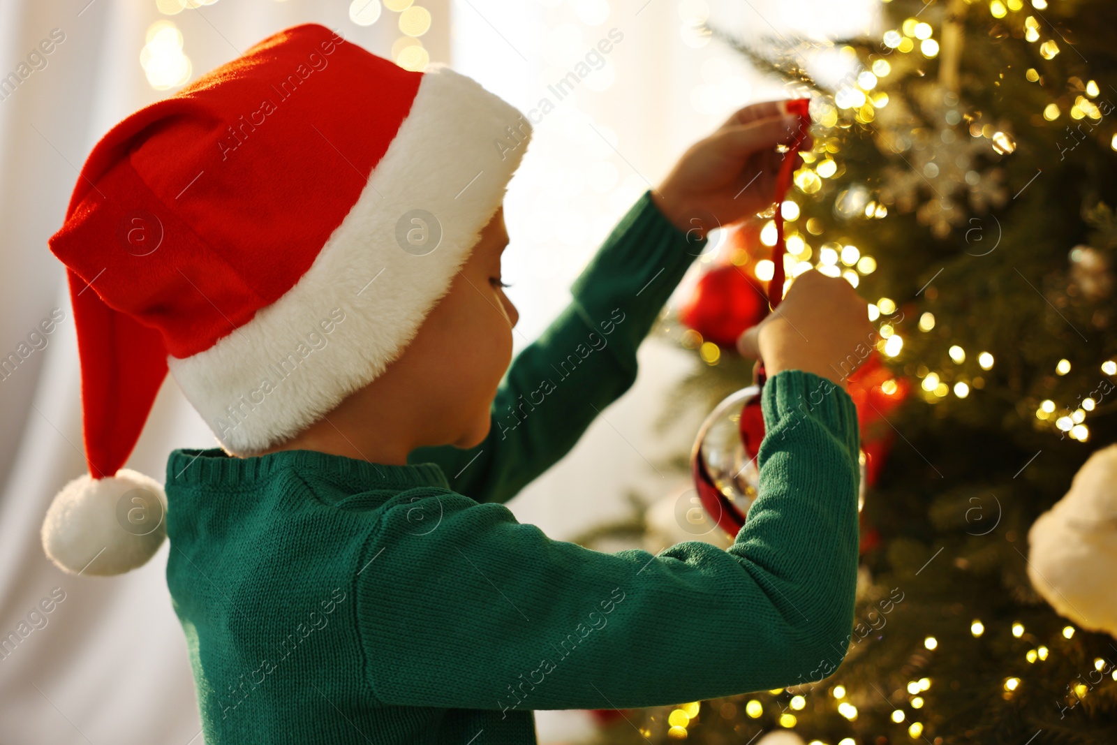 Photo of Cute little boy in Santa hat decorating Christmas tree at home