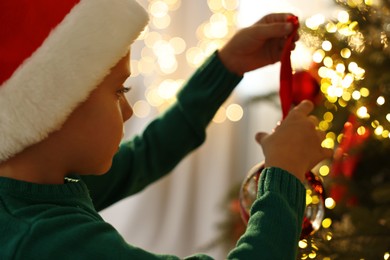 Photo of Cute little boy in Santa hat decorating Christmas tree at home