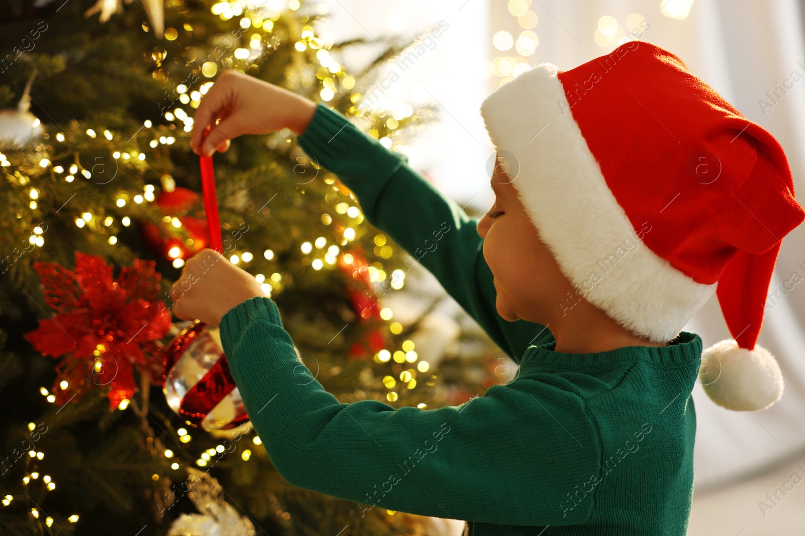 Photo of Cute little boy in Santa hat decorating Christmas tree at home