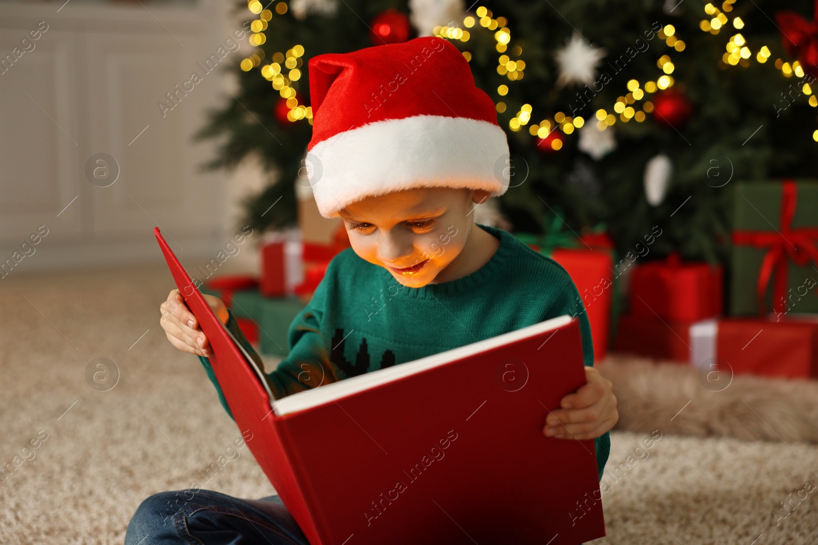 Photo of Cute little boy with Santa hat reading book on rug in room decorated for Christmas