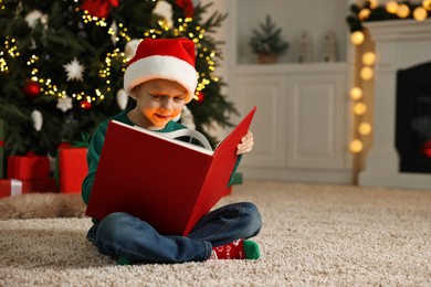 Photo of Cute little boy with Santa hat reading book on rug in room decorated for Christmas, space for text