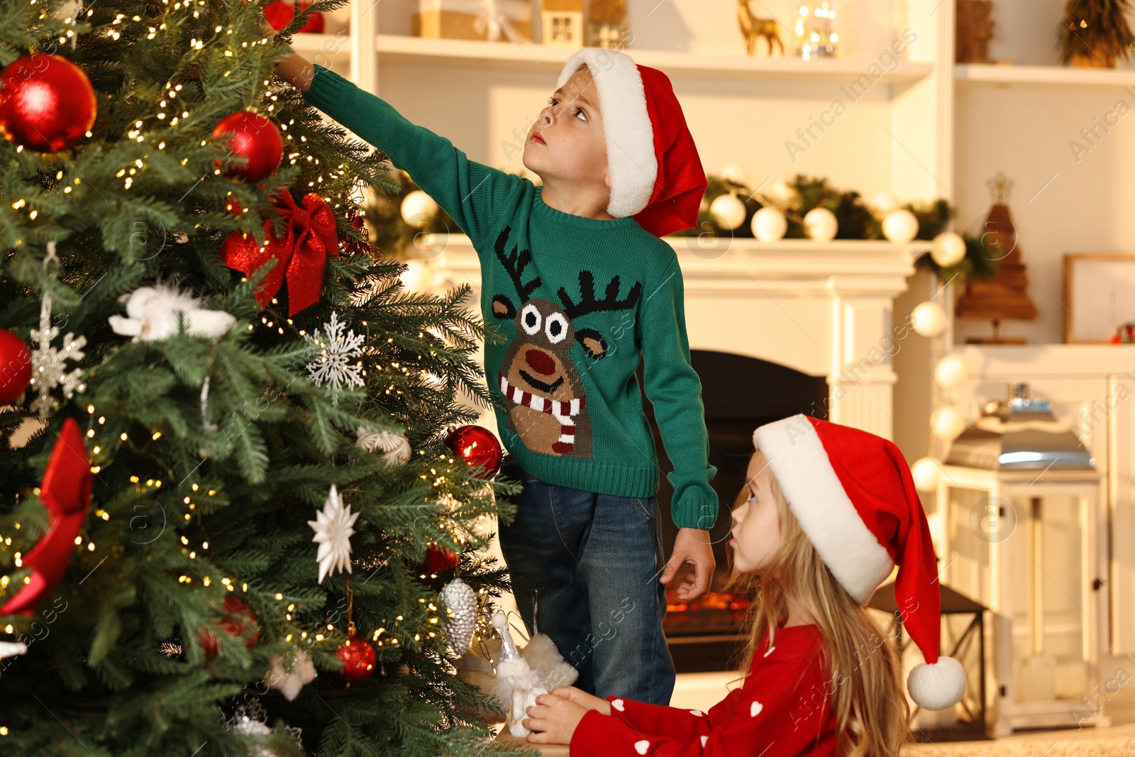 Photo of Little kids in Santa hats decorating Christmas tree at home