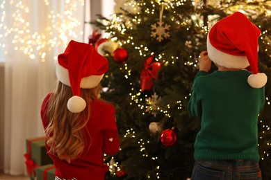 Photo of Little kids in Santa hats decorating Christmas tree at home, back view