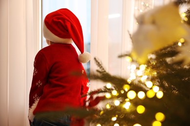 Photo of Little boy with Santa hat near window in room decorated for Christmas, back view