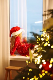 Photo of Little girl with Santa hat near window in room decorated for Christmas