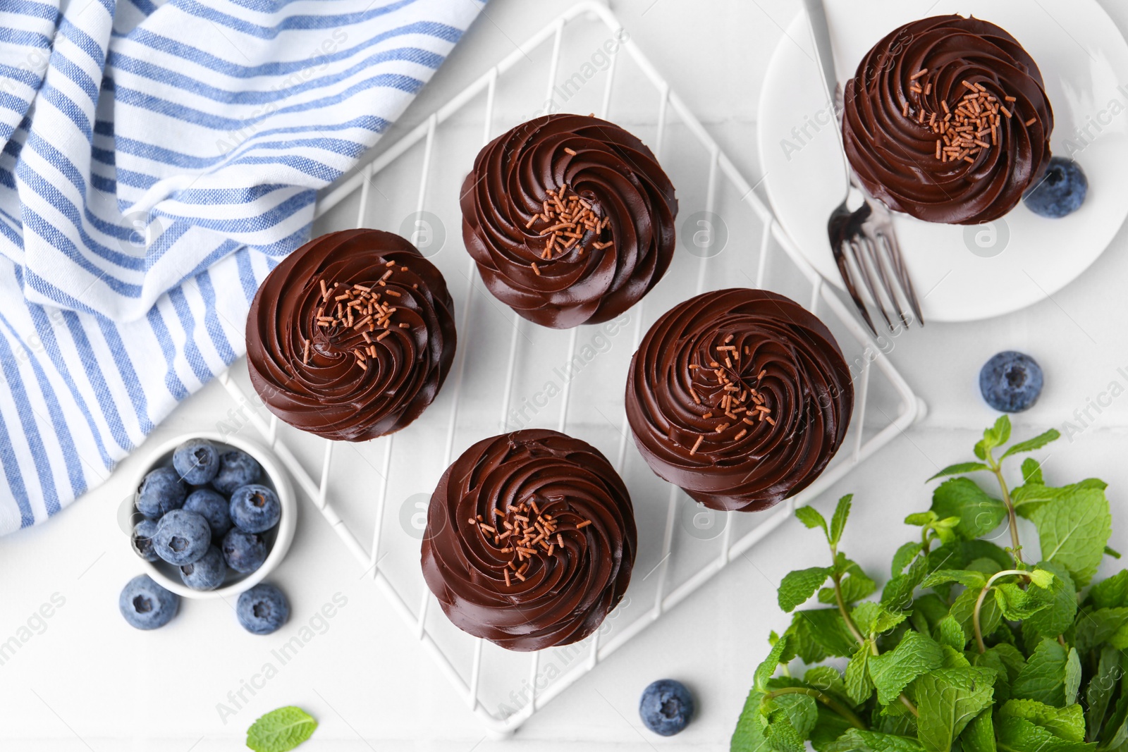 Photo of Tasty cupcakes with chocolate cream and blueberries on white tiled table, flat lay