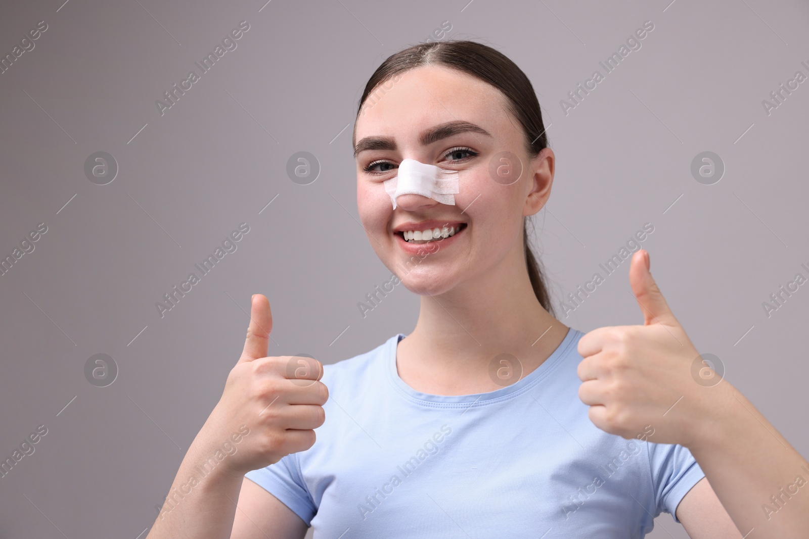 Photo of Woman with medical bandage on her nose after plastic surgery operation showing thumbs up against grey background