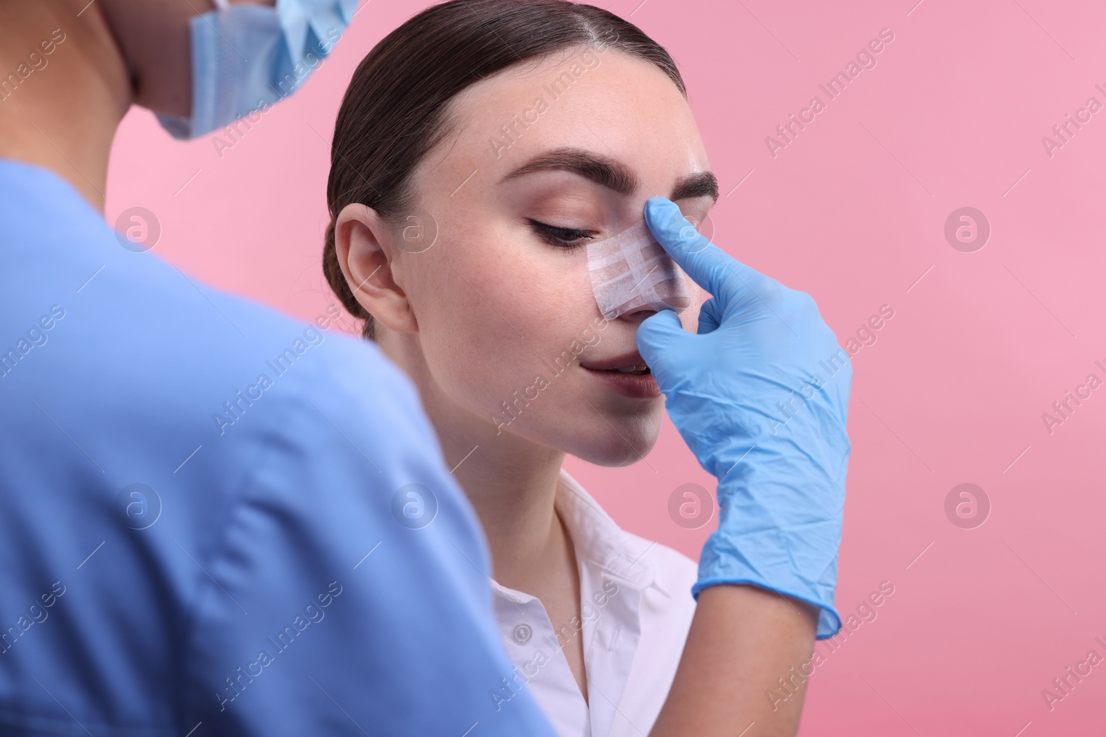 Photo of Doctor checking patient's nose after plastic surgery operation on pink background, closeup