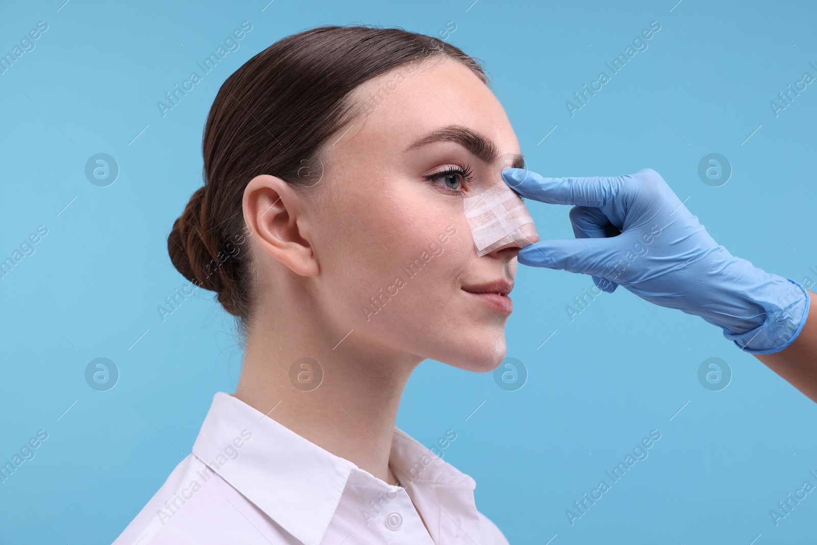 Photo of Doctor checking patient's nose after plastic surgery operation on light blue background, closeup