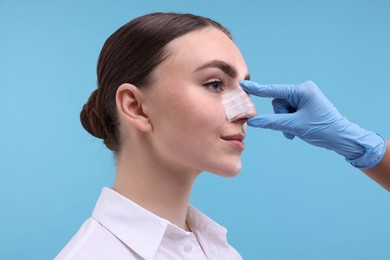 Photo of Doctor checking patient's nose after plastic surgery operation on light blue background, closeup