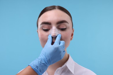 Photo of Doctor checking patient's nose after plastic surgery operation on light blue background, closeup