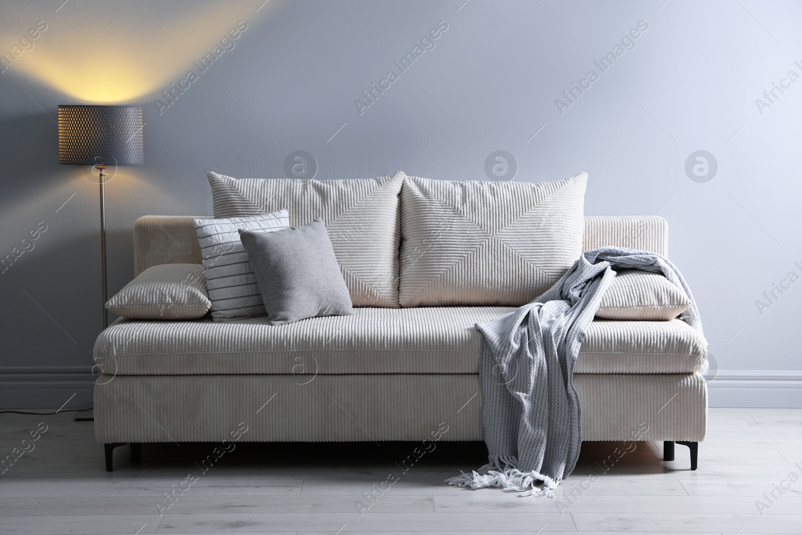 Photo of Comfortable sofa with pillows, blanket and lamp near grey wall indoors