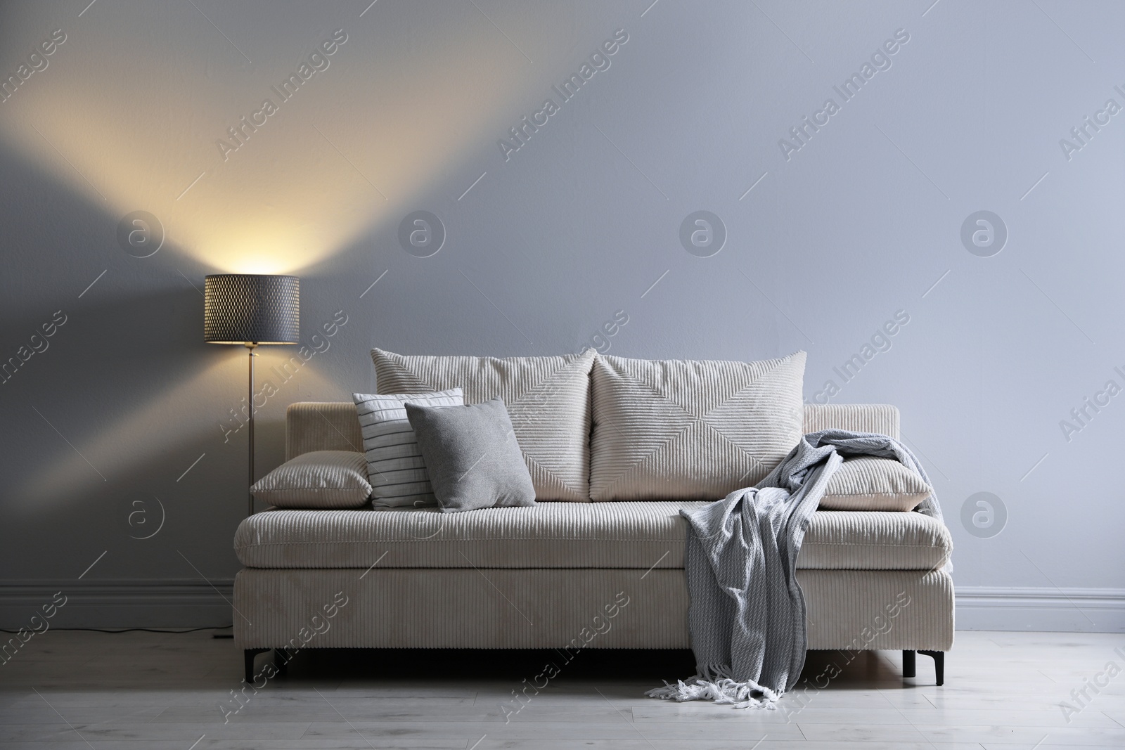 Photo of Comfortable sofa with pillows, blanket and lamp near grey wall indoors