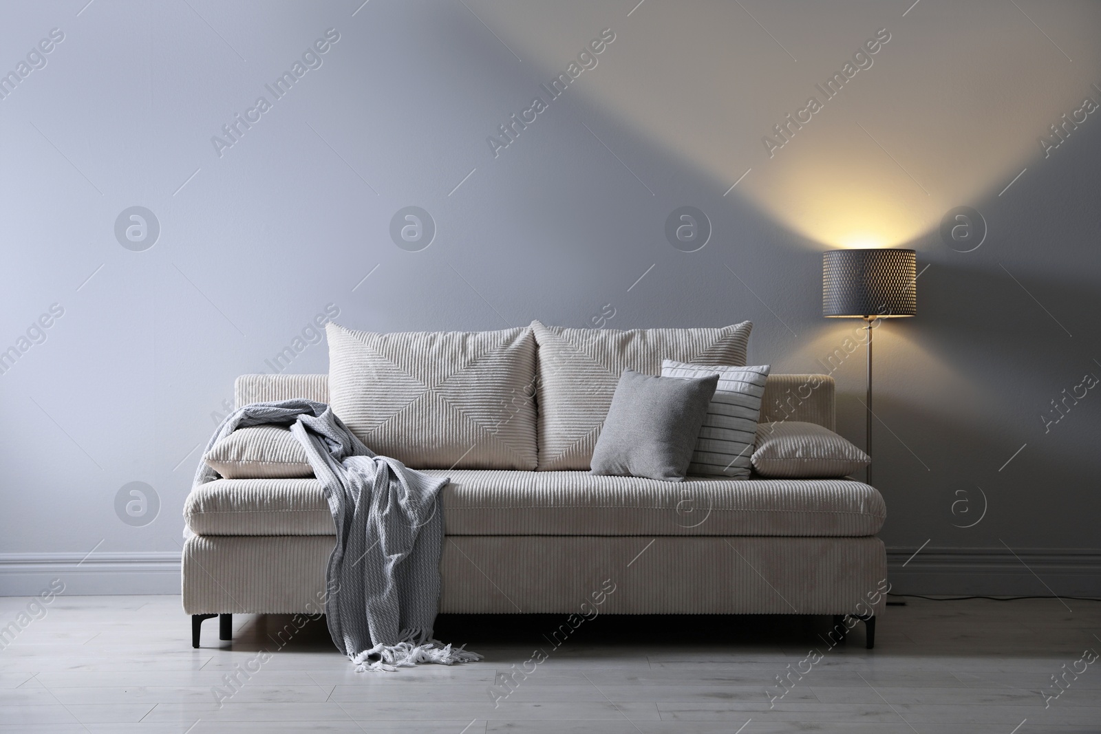 Photo of Comfortable sofa with pillows, blanket and lamp near grey wall indoors