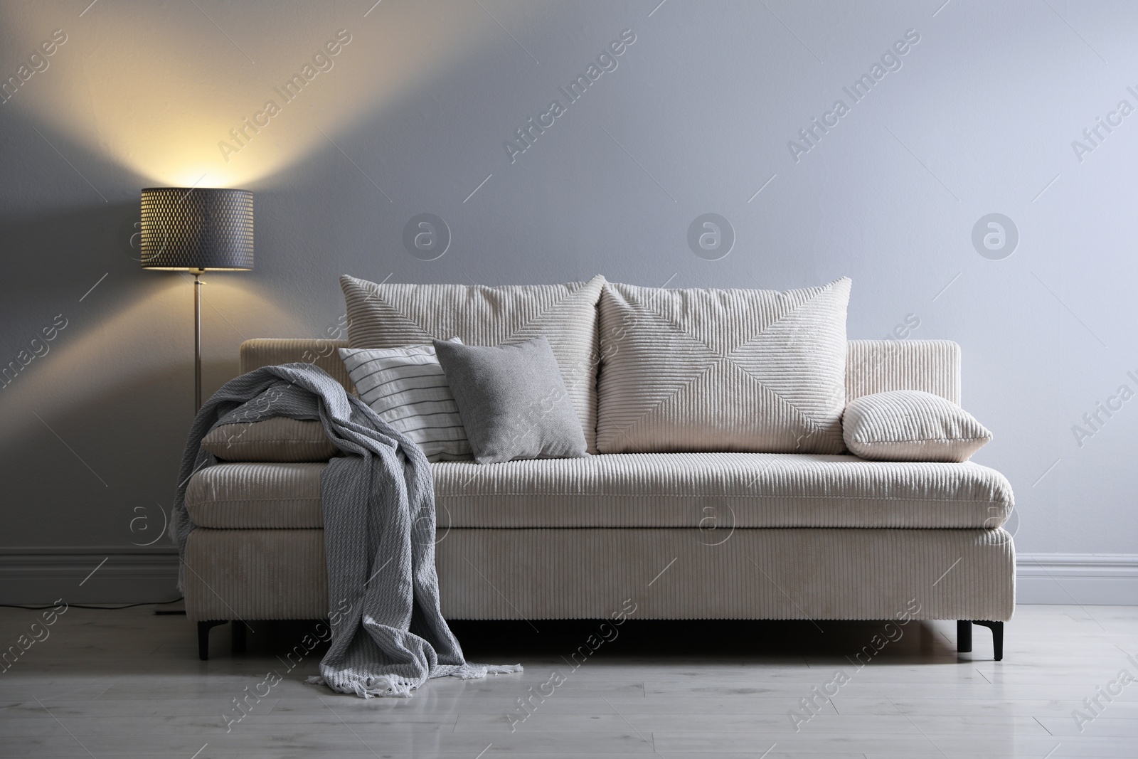 Photo of Comfortable sofa with pillows, blanket and lamp near grey wall indoors