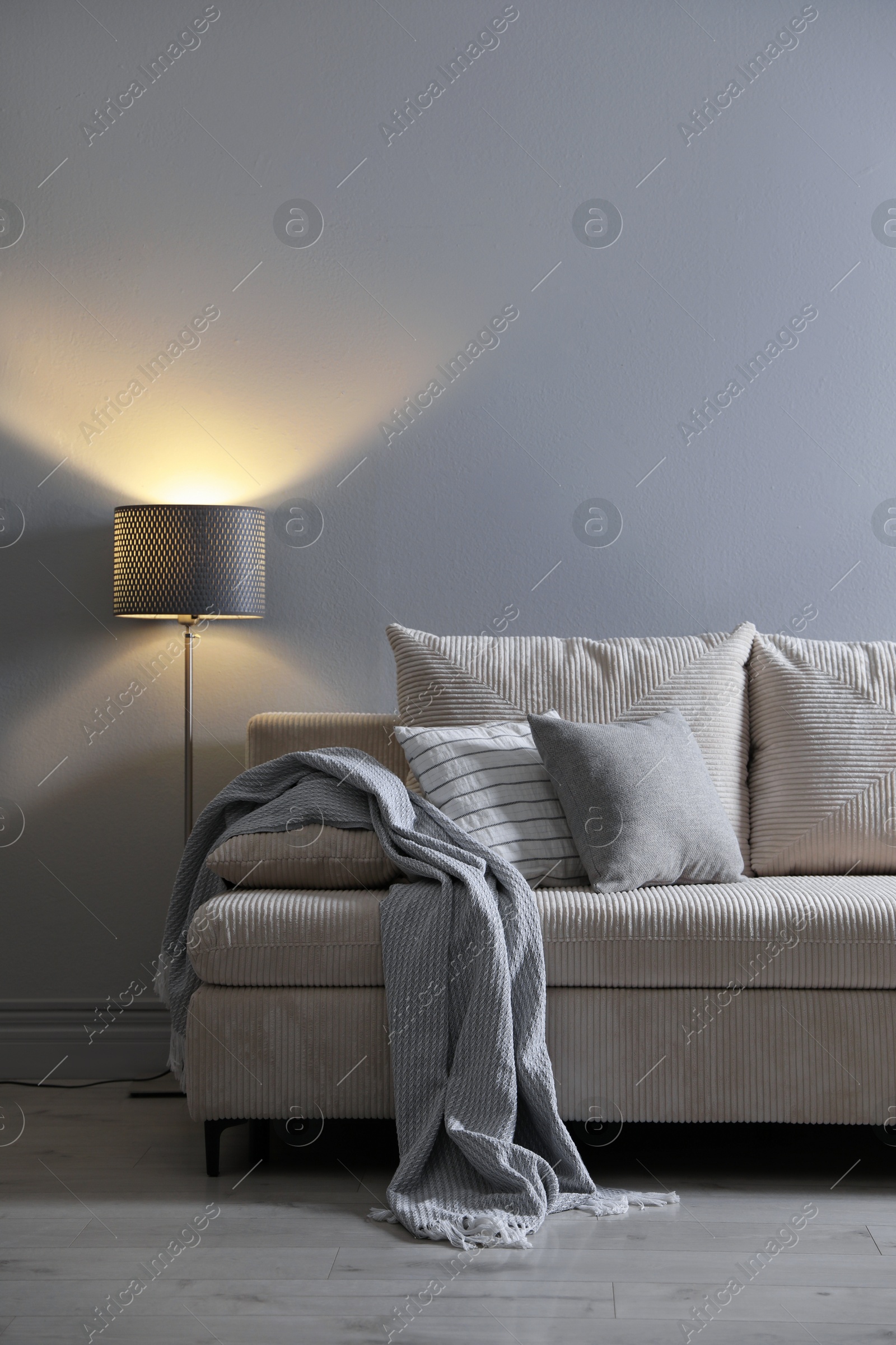 Photo of Comfortable sofa with pillows, blanket and lamp near grey wall indoors