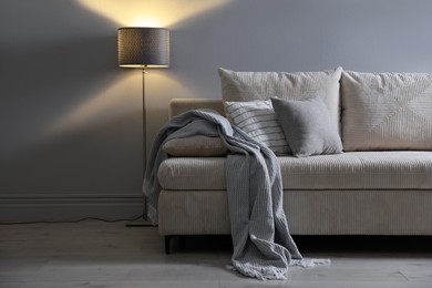 Photo of Comfortable sofa with pillows, blanket and lamp near grey wall indoors