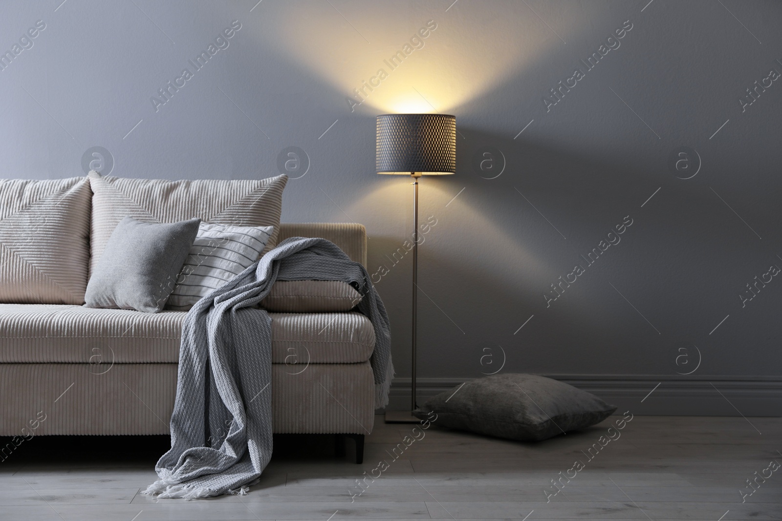 Photo of Comfortable sofa with pillows, blanket and lamp near grey wall indoors