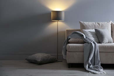 Photo of Comfortable sofa with pillows, blanket and lamp near grey wall indoors