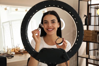 Photo of Beautiful young woman applying face powder with brush indoors, view through ring lamp