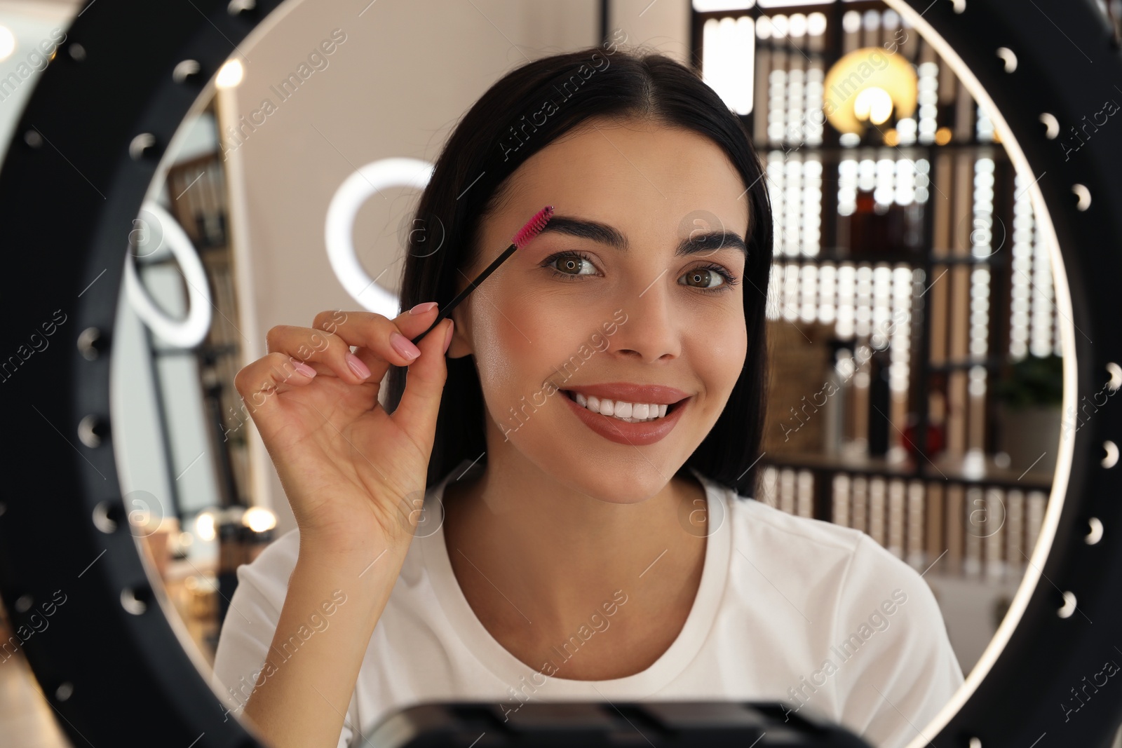 Photo of Beautiful young woman brushing eyebrows indoors, view through ring lamp