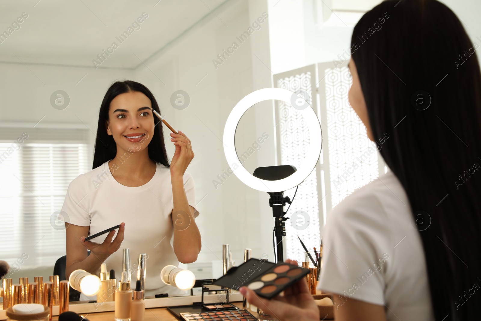 Photo of Beautiful young woman applying makeup at table with mirror and ring lamp