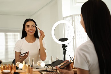 Photo of Beautiful young woman applying makeup at table with mirror and ring lamp