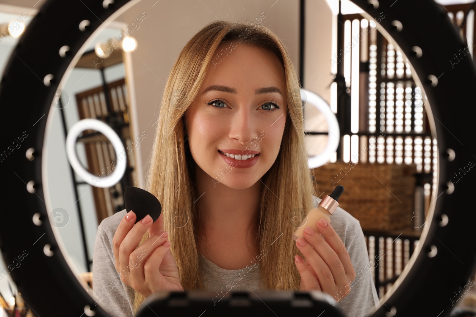 Photo of Beautiful young woman bottle of foundation and beauty blender indoors, view through ring lamp