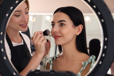 Photo of Professional makeup artist working with beautiful young woman in salon, view through ring lamp