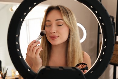 Photo of Beautiful young woman applying blusher with brush indoors, view through ring lamp