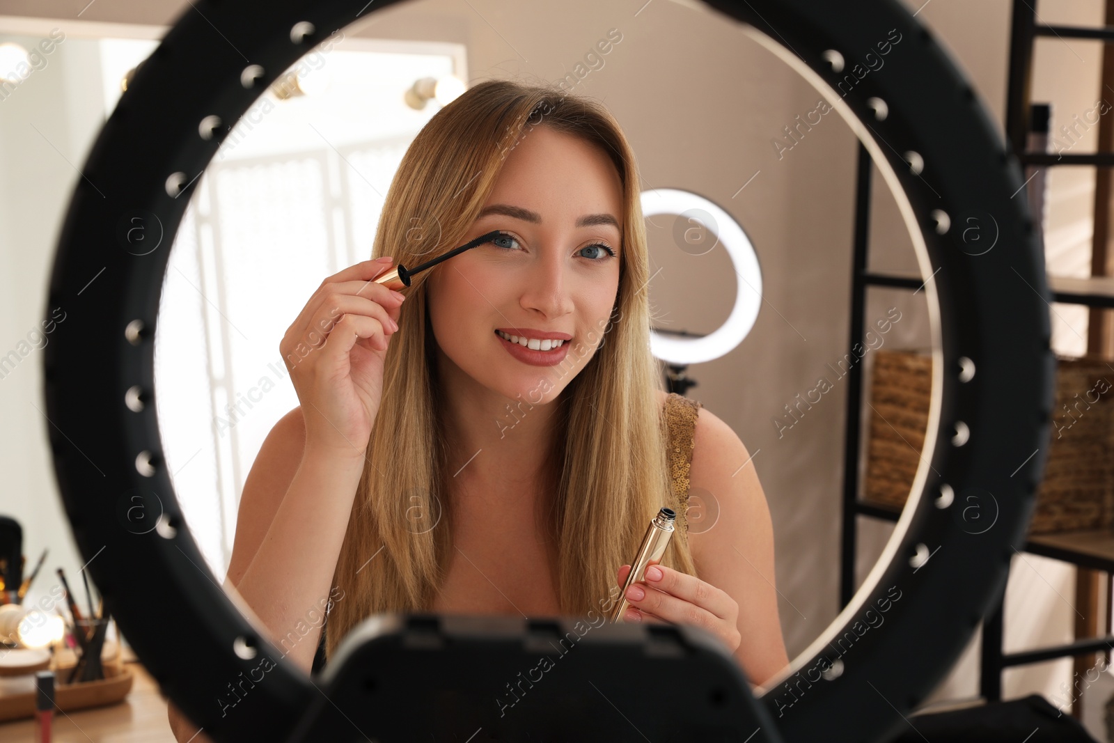 Photo of Beautiful young woman applying mascara indoors, view through ring lamp