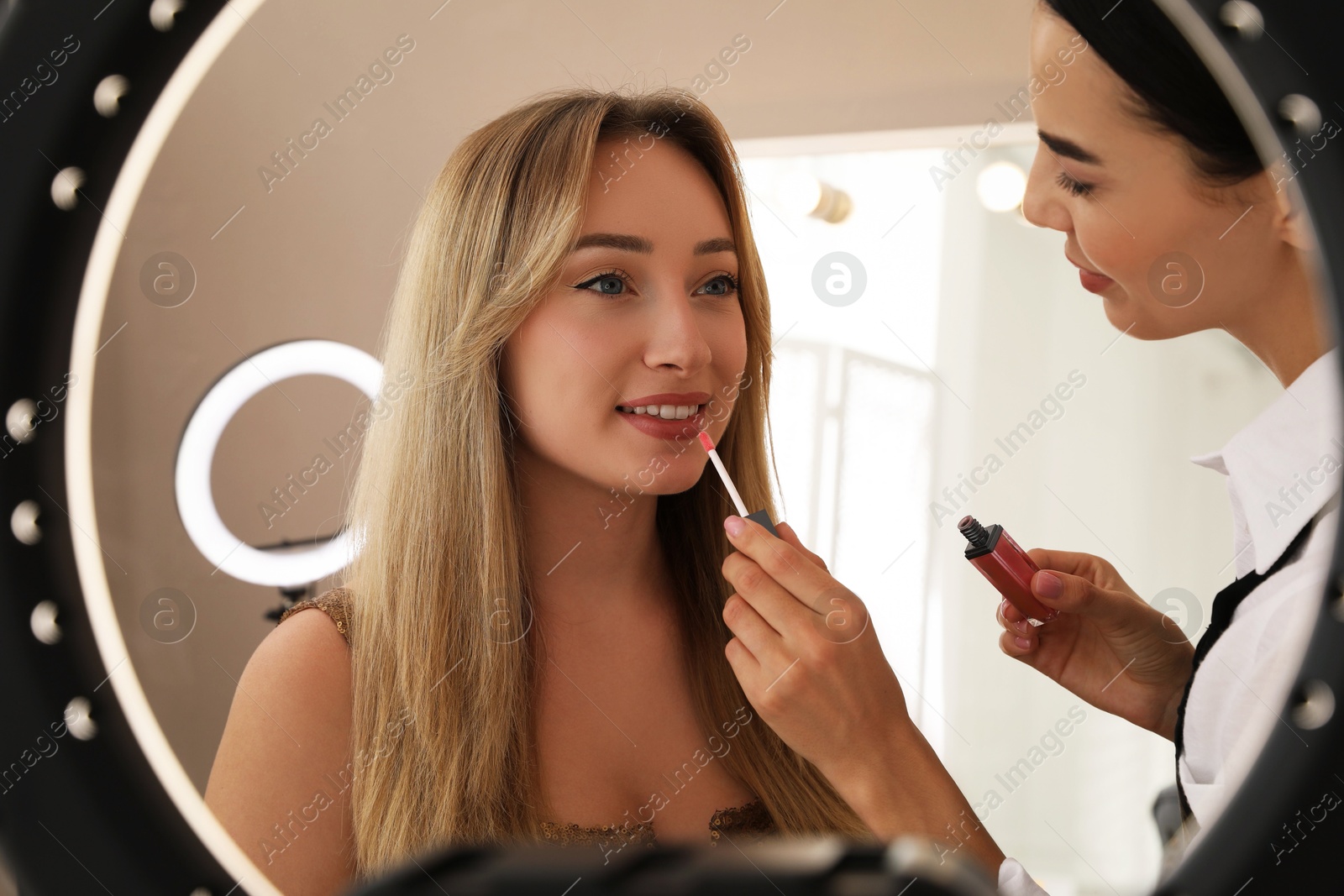 Photo of Professional makeup artist working with beautiful young woman in salon, view through ring lamp