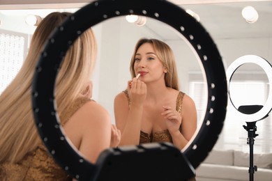 Photo of Beautiful young woman applying lipstick near mirror indoors, view through ring lamp