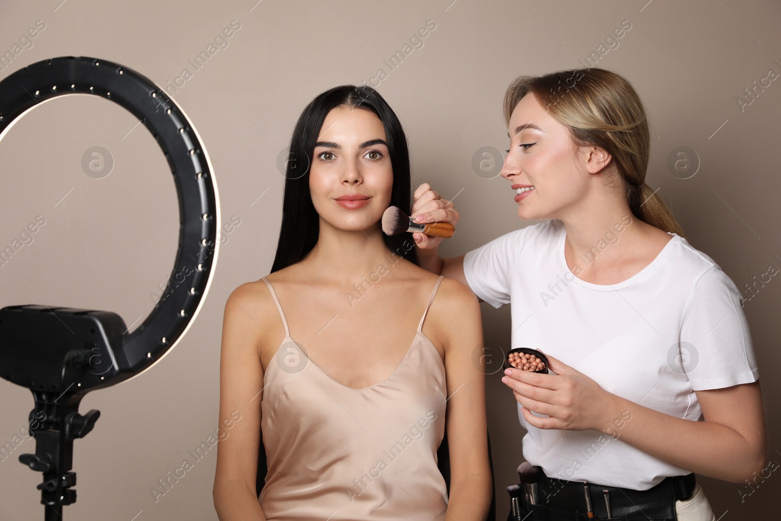 Photo of Professional makeup artist working with beautiful young woman against beige background. Using ring lamp