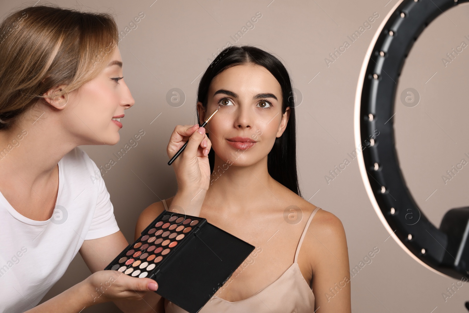 Photo of Professional makeup artist working with beautiful young woman against beige background. Using ring lamp