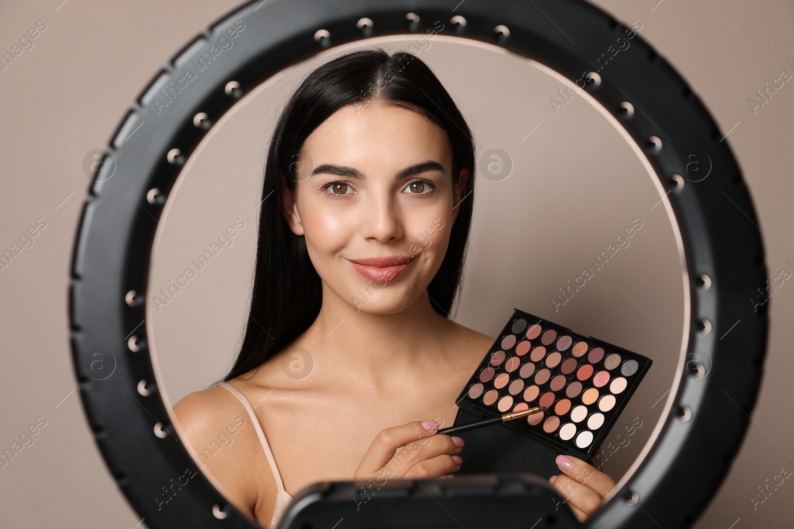 Photo of Beautiful young woman with eye shadow palette on beige background, view through ring lamp