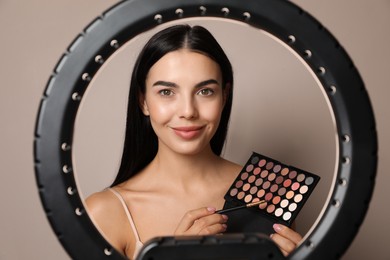 Photo of Beautiful young woman with eye shadow palette on beige background, view through ring lamp