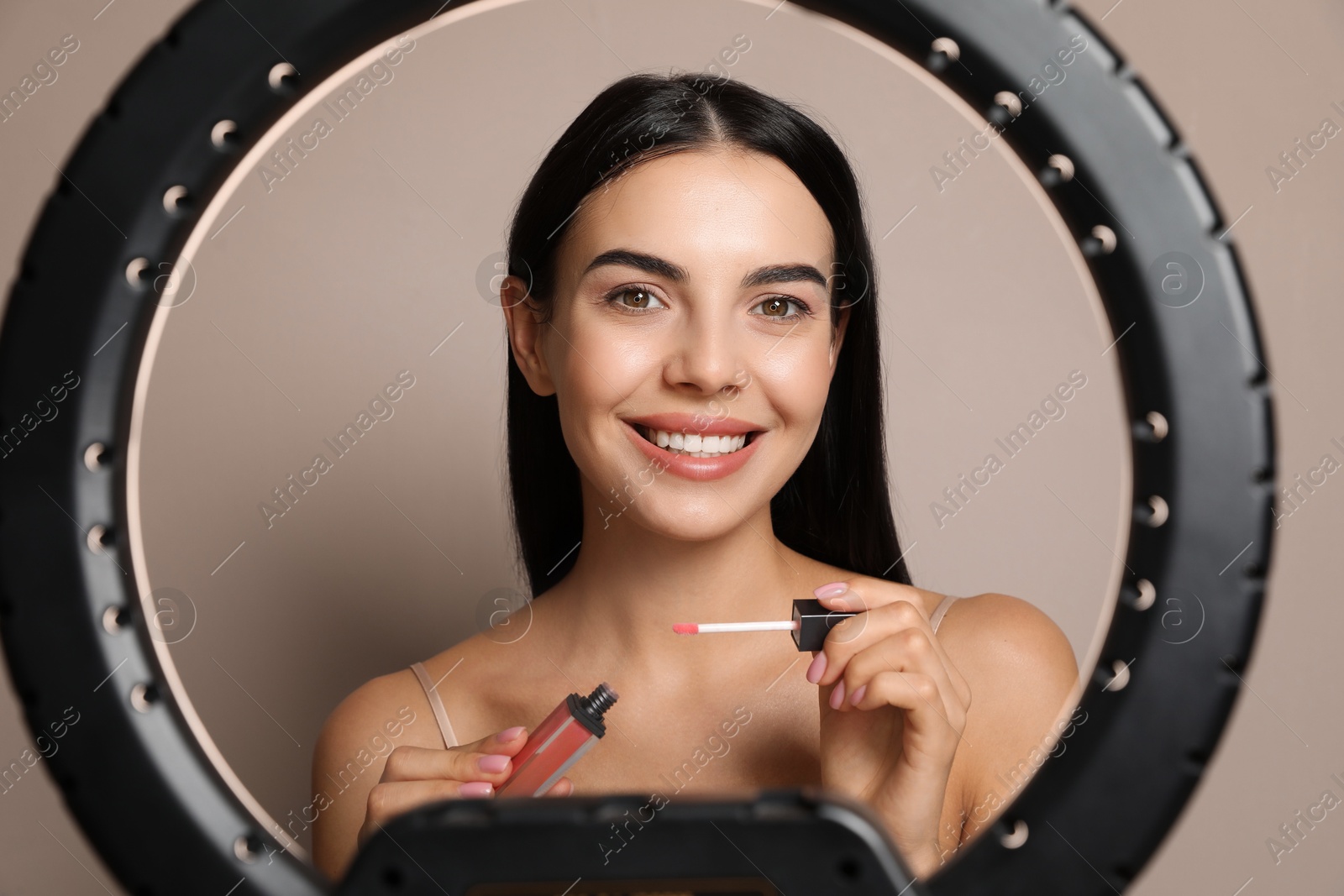 Photo of Beautiful young woman applying liquid lipstick on beige background, view through ring lamp