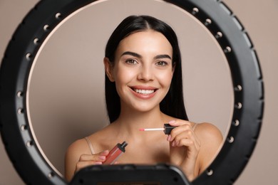 Beautiful young woman applying liquid lipstick on beige background, view through ring lamp