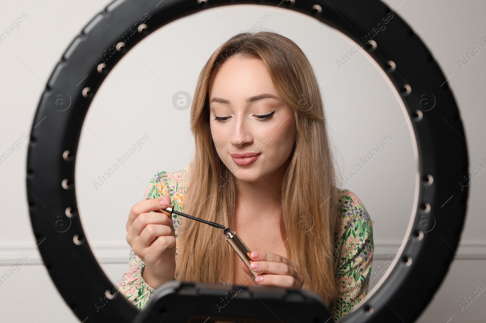 Photo of Beautiful young woman with mascara for eyelashes near white wall, view through ring lamp